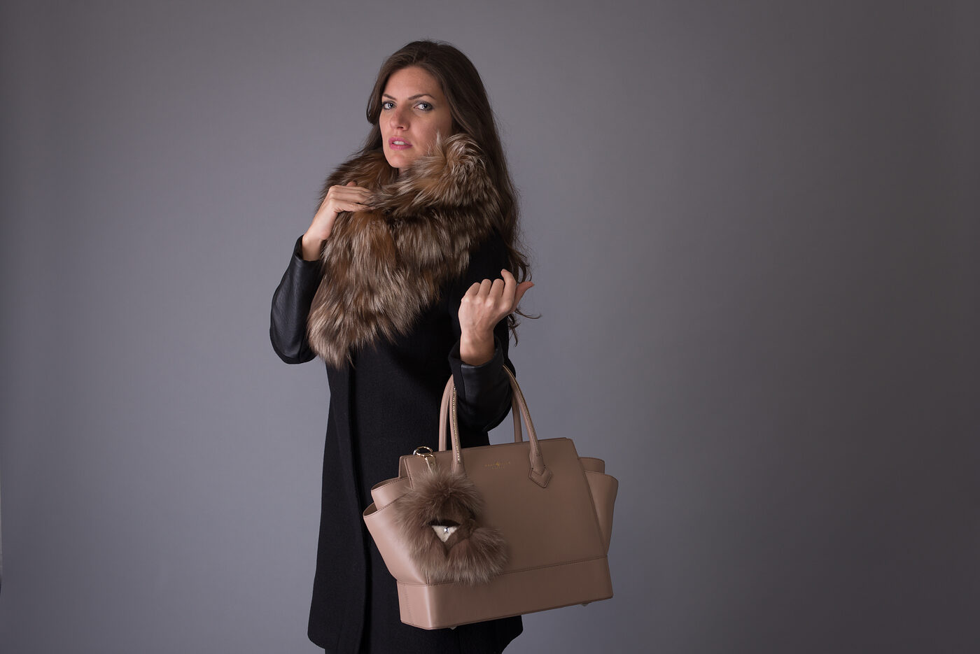 A woman in a black coat with a large natural fox fur collar, holding a taupe leather bag with a fox fur pom-pom