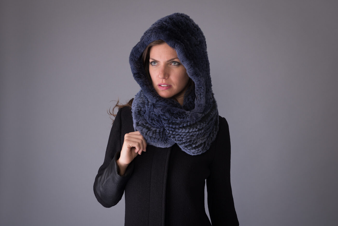 A woman in a knitted blue rex rabbit fur hood and snood, black coat, grey studio backdrop